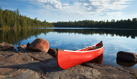 A red canoe resting on a rocky shoreline beside a calm lake, surrounded by lush green trees under a blue sky. Minnesota charter bus travel destination.